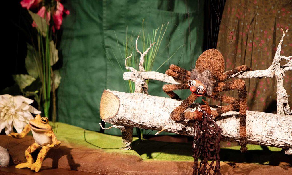 Auf einer Puppenspielbühne sitzt eine große Spinne auf einem Ast und spricht mit einem Frosch.