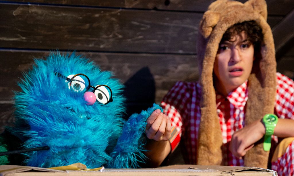 Szene aus Das Klugscheißerchen, ein blaues Plüschmonster sitzt zusammen mit einer Person im Karohemd in einer Hütte, die beiden halten Händchen