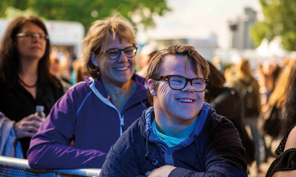 Junge mit Down-Syndrom steht lachend im Publikum, hinter ihm zwei Frauen