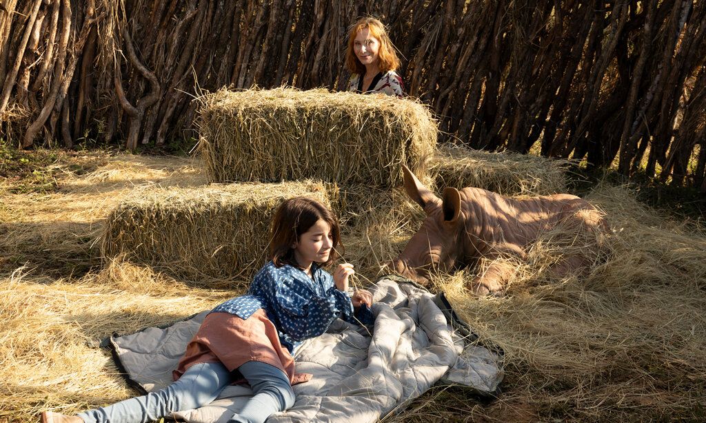 Ein Mädchen liegt mit einem jungen Nashorn im Stroh, im Hintergrund guckt eine Frau hinter einem Strohballen hervor