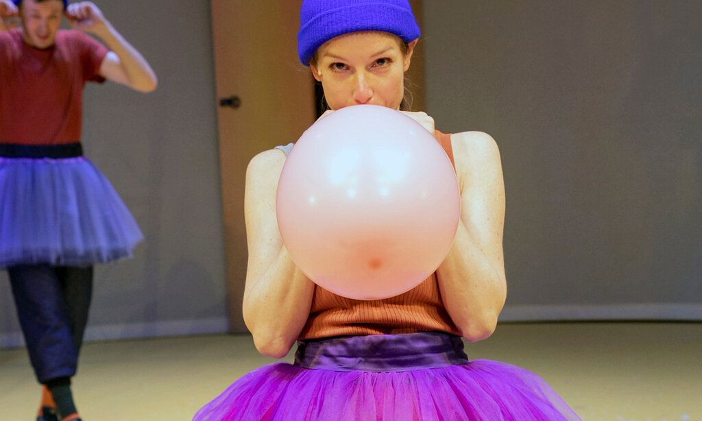 Schauspielerin im Tutu bläst Luftballon auf, Schauspieler im Tutu im Hintergrund hält sich die Ohren zu