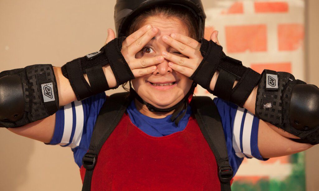Ein Mädchen in Rollschuh- Schutzmontur hält sich die Hände vor ihr Gesicht und blinzelt ängstlich zwischen ihren Fingern hindurch.