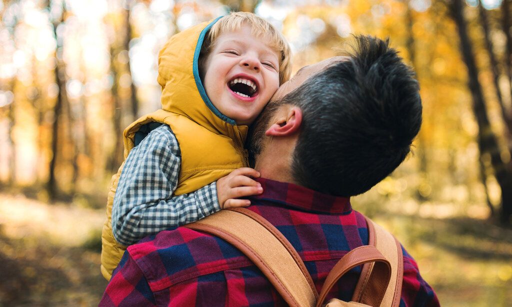 kleiner Junge ist auf dem Arm seines Vaters und  lacht, im Hintergrund herbstlicher Wald