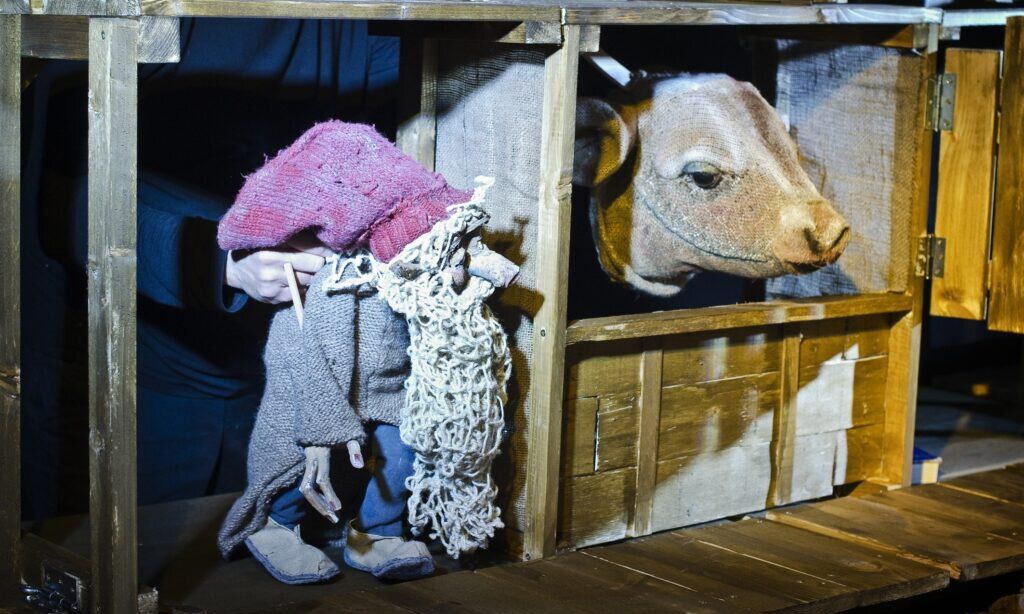 Ein Puppenspieler steht hinter einer Holzkulisse und spielt mit einer Wichtel- und einer Kuhhandpuppe