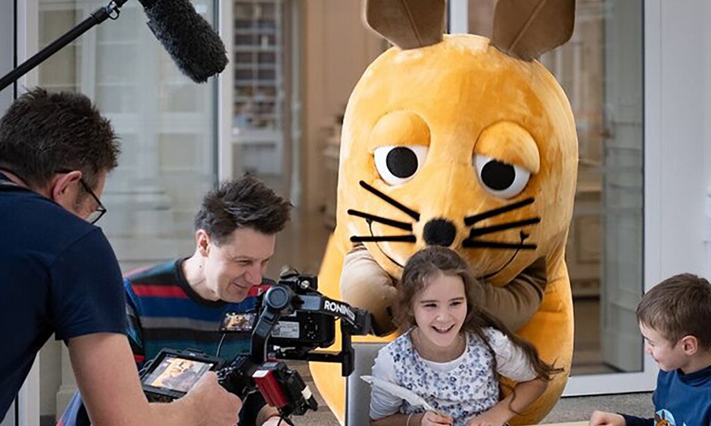 Die Maus steht hinter einem lachenden Mädchen, ein Junge steht daneben und ein Kamerateam ist dabei