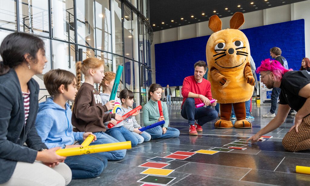 Kinder sitzen mit André Gatzke auf dem Fußboden und unterhalten sich. Die Mais steht daneben. Ein Mann legt bunte Karten auf den Boden.