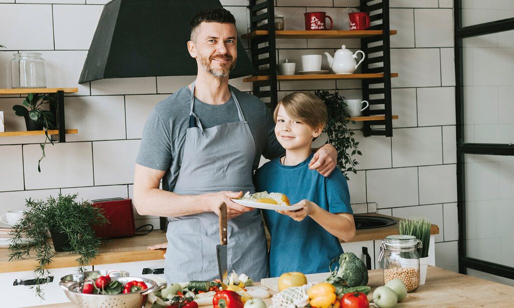Vater und Sohn stehen in der Küche, vor ihnen liegt Gemüse, sie halten zusammen einen Teller, der Vater hat den Arm um den Sohn gelegt