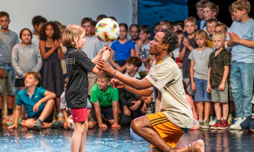 ein Mädchen balanciert einen Fußball mit der Hand, ein Mann hilft ihr dabei, im Hintergrund gucken andere Kinder zu