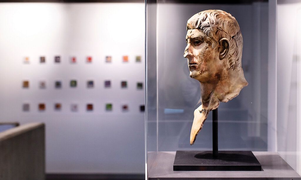 alte römische Büste in einer Glasvitrine, im Hintergrund kleine Bilder an der Wand, unscharf