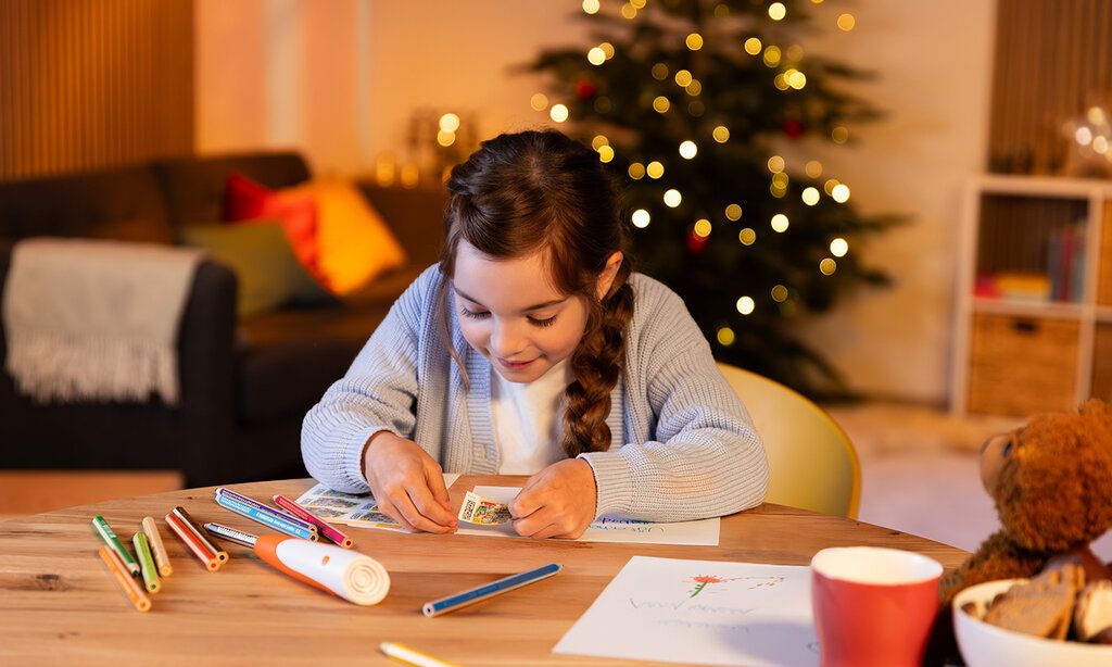 ein Mädchen klebt eine Briefmarke auf ein Papier, im Hintergrund Wohnzimmer mit Weihnachtsbaum