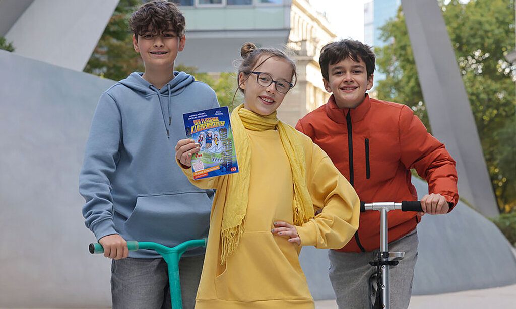 zwei Jungen mit Scootern und ein Mädchen stehen vor einem Gebäude, das Mädchen hält ein Buch in die Kamera