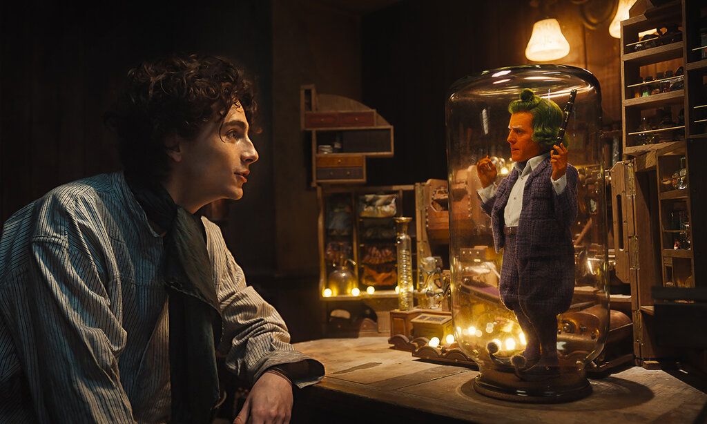 Ein junger Mann sitzt in einem dunklen Zimmer und spricht mit einem sehr kleinen Mann, der auf einem Tisch, unter einer Glasglocke steht