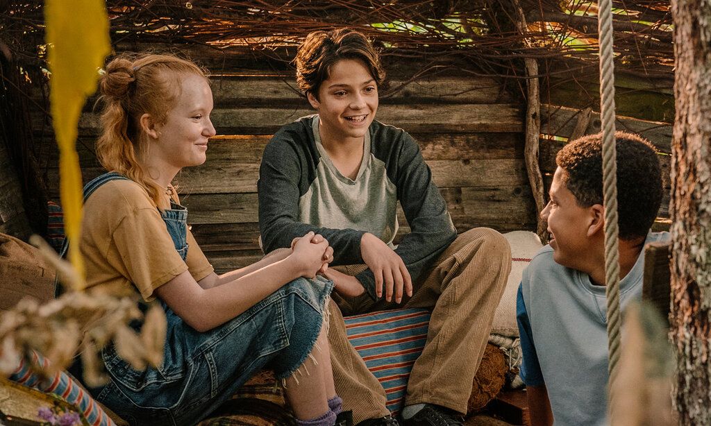 ein Mädchen und zwei Jungen sitzen in einer Holzhütte und unterhalten sich