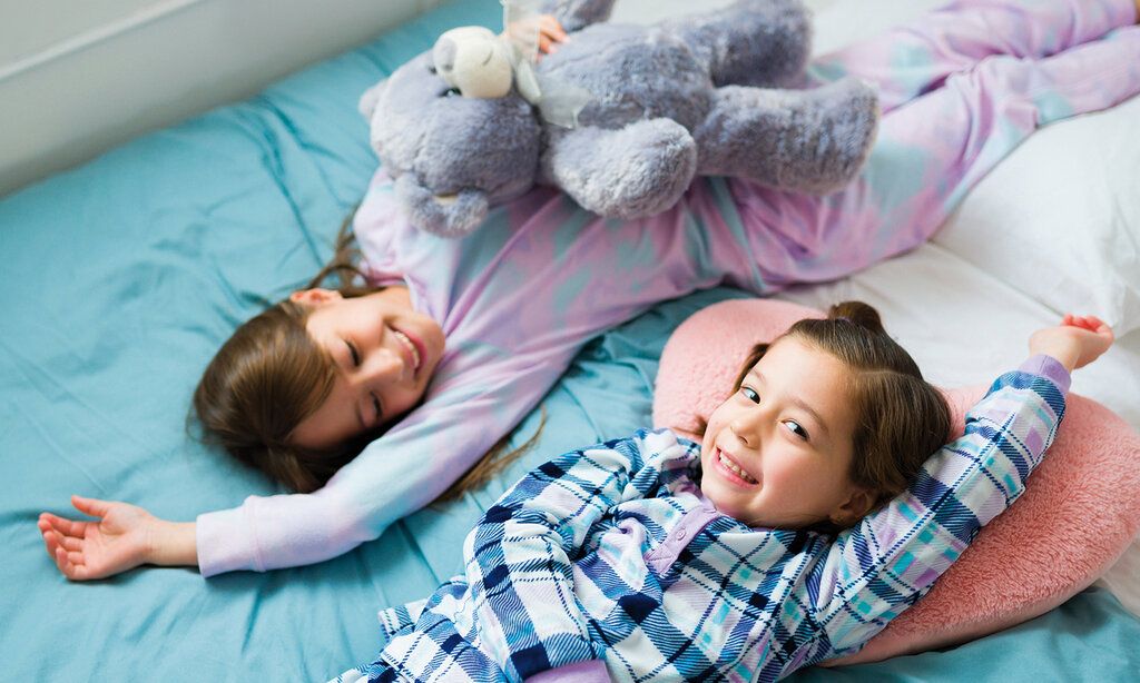 Zwei Mädchen lieben auf einem Bett, in gegengesetzten Richtungen, eine hat ein Plüschtier im Arm