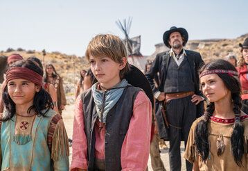 Drei Kinder in Westernkostümen in Prärie im Hintergrund Erwachsene