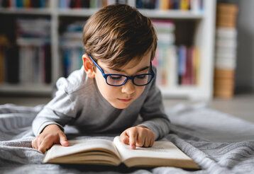 Kleiner Junge mit Brille liegt im Bett undliest ein Buch