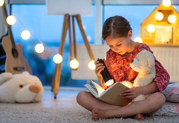 Mädchen sitzt mit einem Teddy auf dem Boden und liest mit einer Taschenlampe in einem Buch, weihnachtliche Stimmung