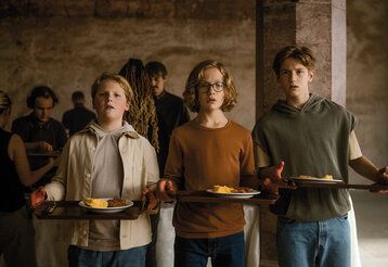 Drei Jungs stehen mit Tabletts, auf denen Teller mit Essen sind, in einem dunklen Gewölbe