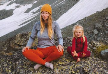 Mutter und Tochter sitzen in karger Berglandschaft im Schneidersitz und machen Yoga
