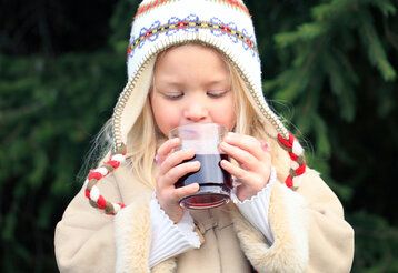 Winterlich gekleidetes Mädchen trinkt einen Kinderpunsch, im Hintergrund Tannen