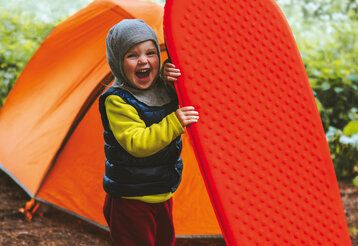 Ein Kind steht vor einem Zelt mit einer Luftmatratze und lacht