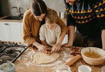 Kleinkind mit Eltern in der Küche, die Mutter hilft ihm dabei, Plätzchen aus Teig auszustechen