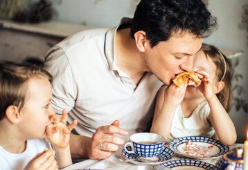 Vater sitzt mit zwei Kindern bei Tisch und beißt in den Pfannkuchen der Tochter