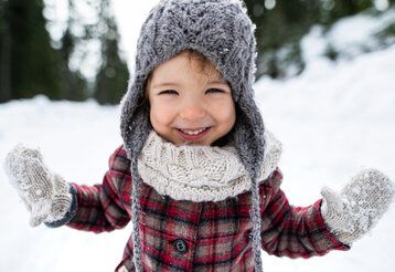 Kleines Kind mit selbstgestrickter Mütze, Handschuhen und Schal im Schnee, lacht in die Kamera
