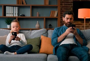 Tochter und Vater sitzen nebeneinander aufm Sofa, beide gucken auf ihr Smartphone