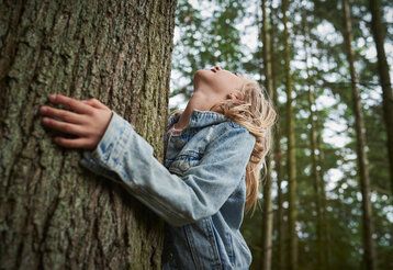 Ein Mädchen in einer Jeansjacke umarmt einen dicken Baumstamm im Wald und schaut dabei nach oben
