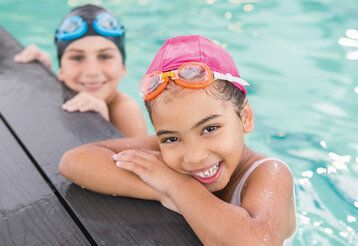 Ein Mädchen mit pinker Badekappe und Chlorbrille mit verschränkten Armen am Schwimmbeckenrand, im Hintergrund ein weiteres Kind