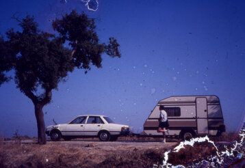 Ein altes Foto von einem Baum, einem Auto mit Wohnwagen und einem Jogger, das Bild ist am rechten Rand zerstört