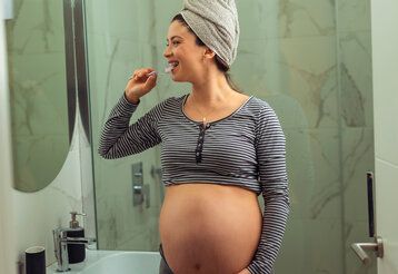 Eine Schwangere steht im Badezimmer, putzt sich die Zähne und schaut dabei in den Spiegel, hellgrüne Kacheln