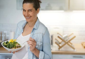 Schwangere Frau steht mit einem Teller, auf dem Brokkoli ist, in der Küche und lächelt in die Kamera