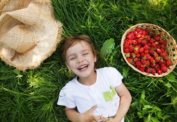 ein Mädchen liegt lachend im Gras, neben ihm ein Sonnenhut und ein Korb mit Erdbeeren, Sicht von oben