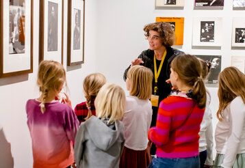 Eine Museumspädagogin steht mit Kindern vor gerahmten Fotos und erklärt diese