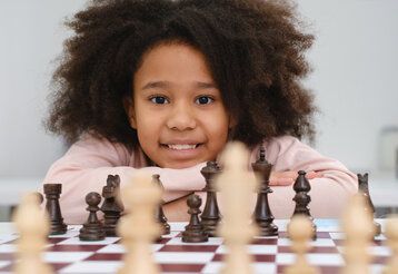 Mädchen hinter Schachbrett, den Kopf auf die verschränkten Arme gestützt, lächelt in die Kamera, im Vordergrund Schachfiguren, teilweise unscharf