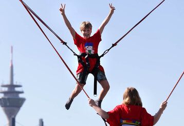 Ein Junge hüpft auf einem Trampolin und macht dabei Victory-Zeichen, im Hintergrund der Rheinturm, davor Mädchen von hinten