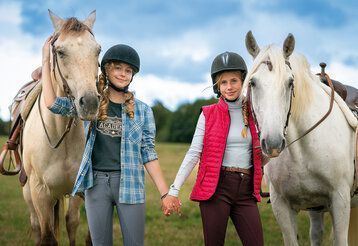 Zwei Mädchen mit Reiterhelmen stehen auf einer Wiese und halten sich an den Händen, neben ihnen jeweils ein Pferd
