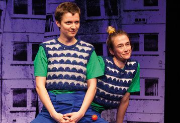 Zwei schauspielerinnen mit gemusterten Pullundern vor violetter Häuderfassaden-Kulisse
