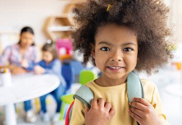 Porträt eines kleinen Schulmädchens, dahinter sitzt eine Lehrerin mit einer weiteren Schülerin an einem Tisch, Hintergrund unscharf