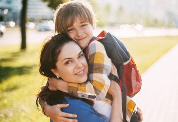 Mutter und Junge mit Schulrucksack umarmen sich, im Hintergrund Straßenszene, Baum und Grünstreifen, unscharf