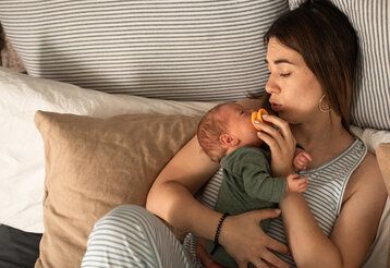 Eine Frau sitzt im Bett mit vielen Kissen und hält ihr neugeborenes Baby im Arm