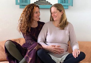 Zwei Frauen sitzen im Schneidersitz nebeneinander, die eine ist schwanger und hält ihre Hand auf ihren Bauch, die andere Frau hält auch ihre Hand auf den Bauch und lächtelt die Schwangere an