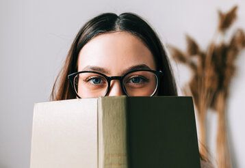 Frau mit Brille hält sich ein Buch vors Gesicht und guckt darüber in die Kamera