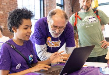 Junge mit violettem T-Shirt arbeitet an einem Laptop, ein älterer Mann im violetten T-Shirt steht neben ihm und spricht mit ihm, ein anderen Mann steht daneben und schaut zu