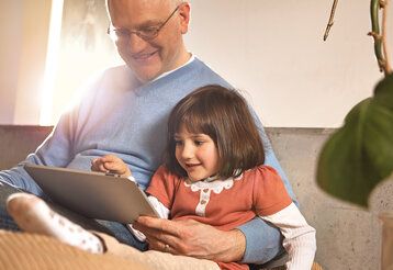 Opa sitzt mit Enkelin zusammen und beide schauen aufs Tablet.