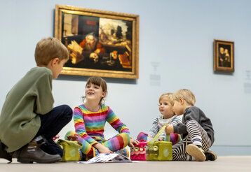vier Kinder sitzen mit Audioguides auf dem Boden im Kunstpalast, im Hintergrund Bilder alter Meister