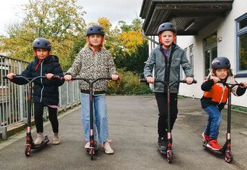 Vier Kinder mit Helmen auf Rollern, schauen in die Kamera
