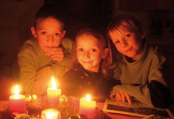 drei Kinder sitzen zusammen, vor ihnen brennende Kerzen vom Adventskranz, dunkle gemütliche Atmosphäre